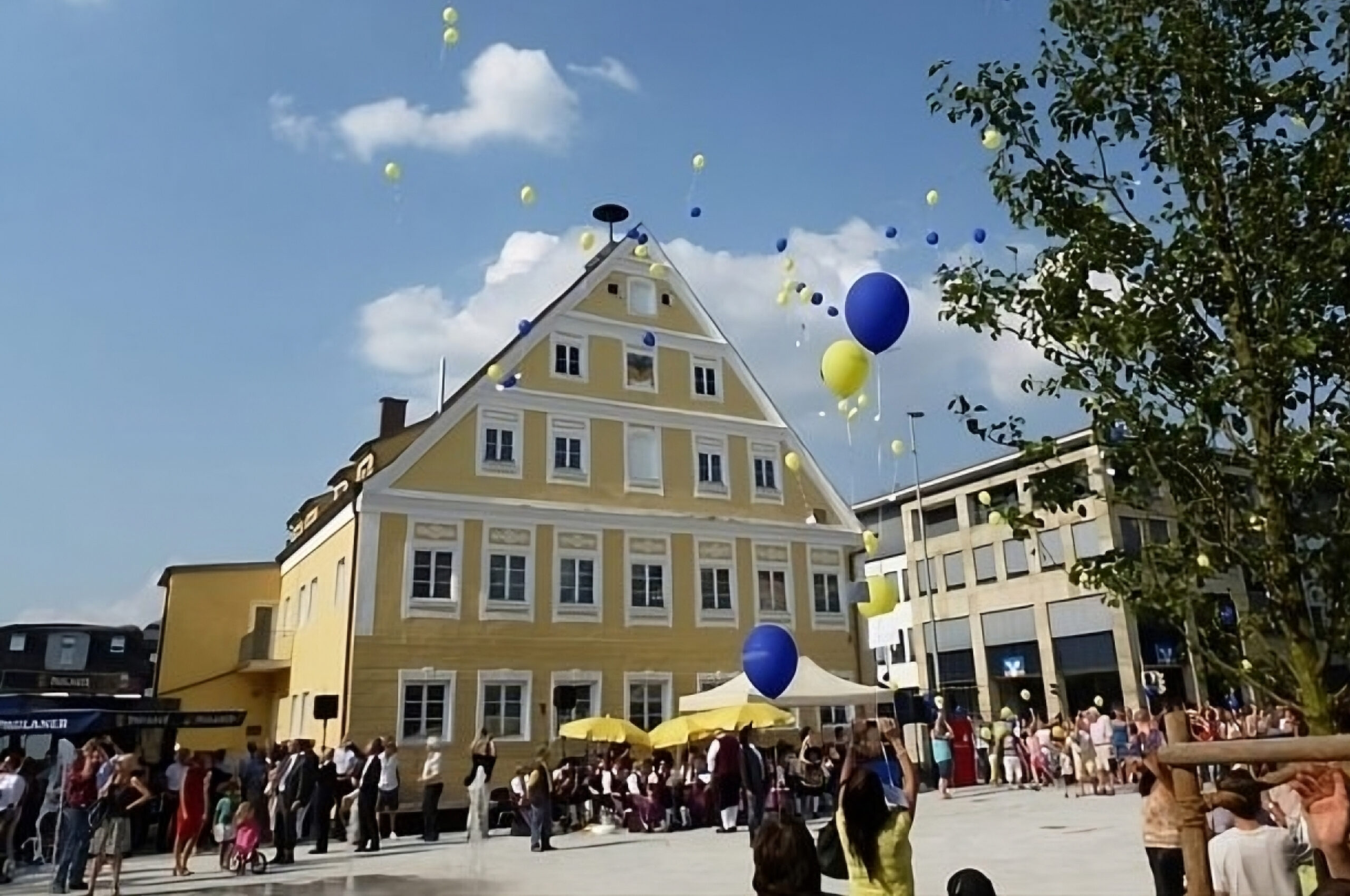 Gelbes Haus auf einem Platz, vor welchem Luftballons aufsteigen.